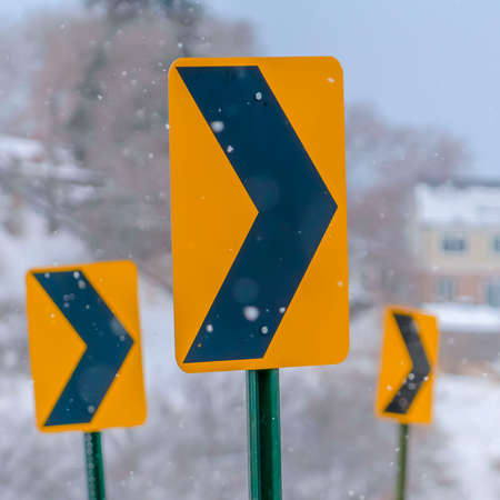 Square Directional road signs against a snowy landscape. Close up of a yellow and black directional road sign in winter. A snow covered landscape with homes and trees can be seen in the hazy background.の写真素材