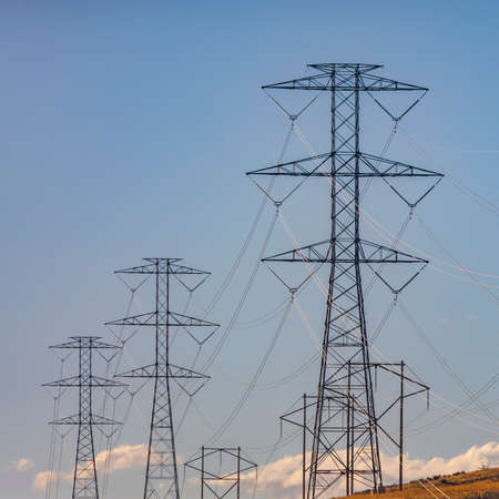 Row of tall utility towers against pale blue sky with puffy clouds. The metal towers are supporting...