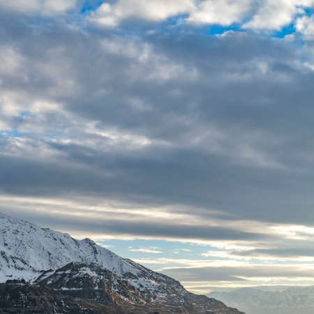 Clear Square Vivid blue sky filled with thick gray clouds over an immense mountain. The rugged...