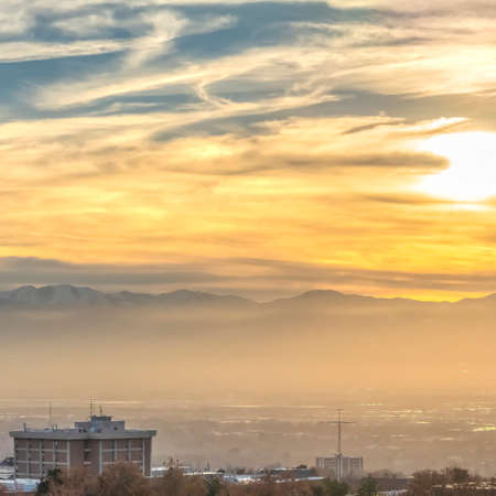 Square Panorama of a bustling city against a towering snowy mountain at sunsetの写真素材