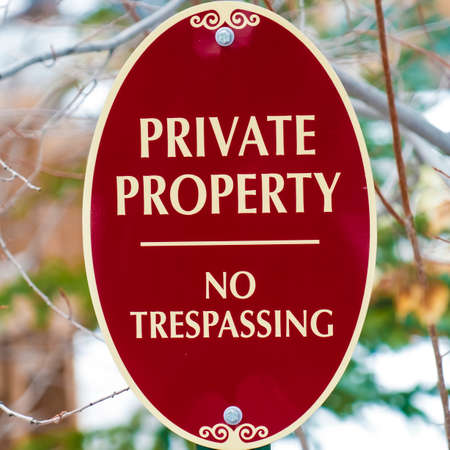 Square Oval shape Private Property No Trespassing sign with red and white colors. Slim branches of a tree on a snowy landscape can be seen behind the sign.の写真素材