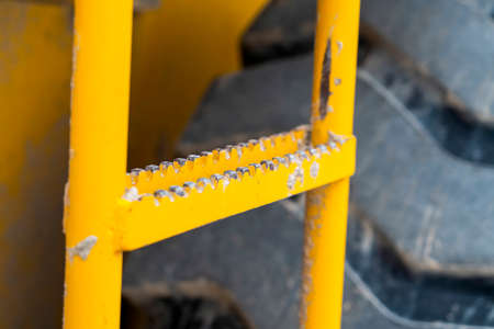 Close up of the ladder with peeling yellow paint of a construction machinery. The huge and heavy-duty black rubber tire of the vehicle can be seen behind the ladder.の写真素材