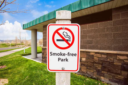 Close up view of a Smoke Free Park sign agianst a building with stone wall. Road, grassy terrain, and blue sky with clouds can also be seen in the background.の写真素材