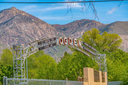 Welcome arch in Ogden Utah against lush trees towering mountain and blue sky. A chain link and concrete fence can be seen in the foreground.の写真素材