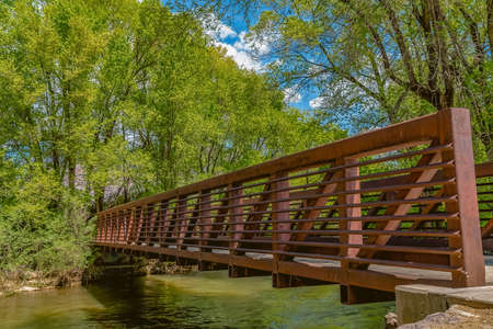 Bridge with latticed metal guardrail over a calm stream viewed on a sunny day. Trees with lush green leaves can be seen on the shoreline.の写真素材
