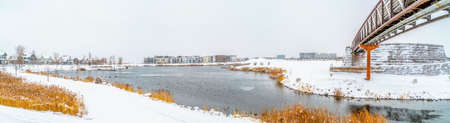 Lake and buildings surrounded with snowy ground under cloudy sky in winter. A circular pathway leads...