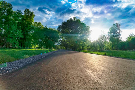 Close up of a paved road illuminated by the bright sun in the cloudy blue sky. A scenic view of vast green field and lush trees can be seen from the road.の写真素材
