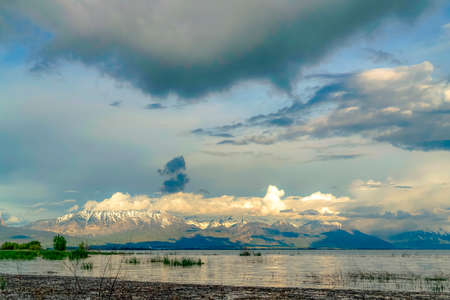 Picturesque view of shiny lake and snow topped mountain under cloudy sky. The bright setting sun is...