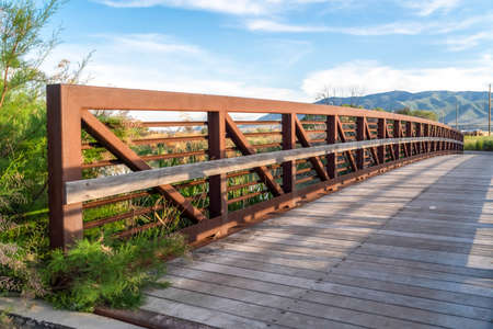Bridge over a lake with view of mountain and valley under blue sky and clouds. The sunlit structure has a wood deck and metal lattice guardrail .の写真素材