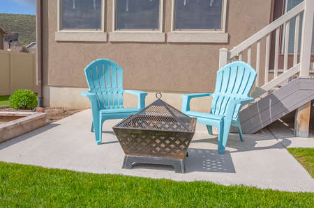 Square fire pit and blue plastic chairs on the yard of a home on a sunny day. Outdoor stairs leading...
