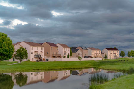 Beautiful homes with a scenic view of a shiny pond and expansive grassy field. Sky filled with thick gray clouds can be seen over the landscape.の写真素材
