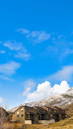 Vertical frame Row of houses on a mountain slope with cloudy blue sky in the background. A mountain peak covered with fresh white snow can also be seen behind the homes.の写真素材