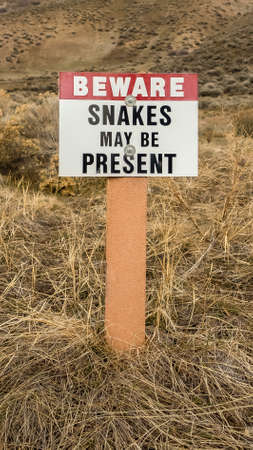 Vertical tall Sign that reads Beware Snakes May Be Present on a grassy mountain. In the background is a view of bright blue sky and snowy mountain peak.の写真素材