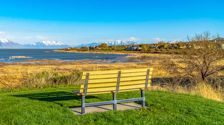 Panorama frame Empty bench overlooking a lake and casting shadow on the field on a sunny day. Lakefront homes and snowy mountain can also be seen under the blue sky.の写真素材