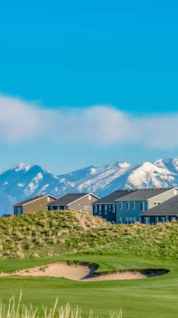 Vertical frame Hill with lush green grasses and houses against blue sky on a sunny day. The striking mountain in the background is covered with snow.の写真素材