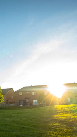 Vertical Sun in the vast blue sky shining brightly over beautiful multi storey houses. The residences have a view of an expansive terrain covered with vibrant green grasses.の写真素材