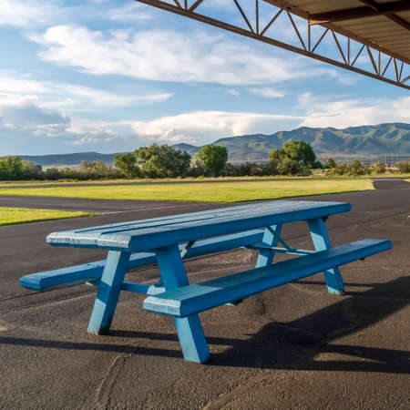 Square Picnic area with spool tabled and blue wooden tables under a pavilion. The park has a scenic view of grassy field, lush trees, mountain, and cloudy sky.の写真素材