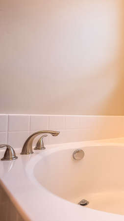 Vertical Close up of a white oval bathtub with stainless steel faucet inside a bathroom. The room has a beige wall and sliding window with blinds.の写真素材