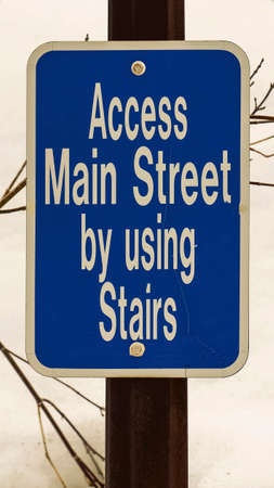 Vertical Close up of a blue sign that reads Access Main Street By Using Stairs. Leafless branches and snow covered ground can be seen in the background.の写真素材
