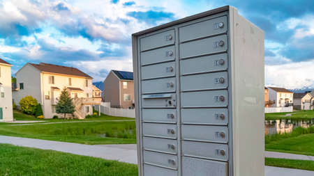 Panorama White metal cluster mailbox with pond grassy terrain and homes in the background. Cloudy sky and snow capped mountain can also be seen in this landscape.の写真素材