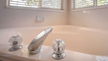 Panorama Polished bathtub with shiny faucet and double crystal handle inside a bathroom. Trees and bright sky can be seen through the windows with blinds.の写真素材