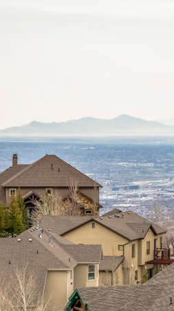 Vertical Rooftops of homes with cloudy sky mountain and vast valley in the background. Vivid green conifers and trees with leafless branches can be seen around the houses.の写真素材