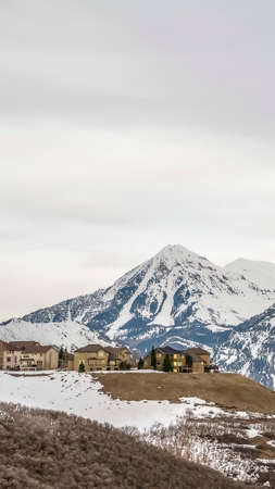 Vertical Hill with homes and snow overlooking a striking white peaked mountain in winter. The sky over the scenic landscape is filled with thick clouds.の写真素材