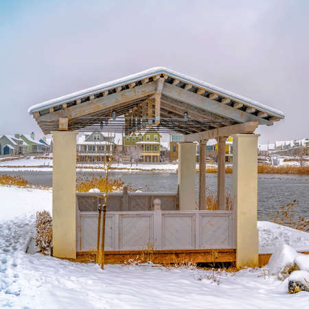 Square frame Pavilion on the snow covered shore of a lake viewed on a cloudy winter day. Yellow grasses, decks, and houses can also be seen around the silvery lake.の写真素材