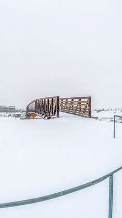 Vertical frame Footprints on the snowy pathway with railings leading to the bridge over a lake. Residential area against cloudy sky in winter can be seen in the distance.の写真素材