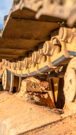 Vertical Close up of the dirty track pad and rollers of a yellow excavator on a sunny day. The construction vehicle is parked on sunlit ground under blue sky.の写真素材