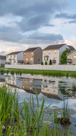 Vertical Beautiful multi storey homes built in front of a grassy and shiny pond. Sky with gray and puffy clouds can be seen over the scenic landscape.の写真素材