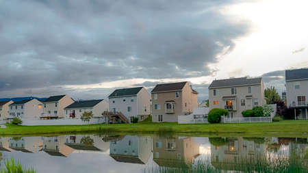 Panorama frame Family homes and bright cloudy sky reflected on the shiny neighborhood pond. The expansive terrain is covered with vivid green grasses.の写真素材