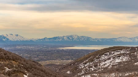 Panorama Lake and vast valley viewed from a mountain with trees and snow. Cloudy sky and snow peaked mountain can also be seen in the distance.の写真素材
