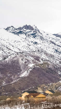 Vertical Residential area at the base of a towering snowy mountain against cloudy sky. Conifers and...