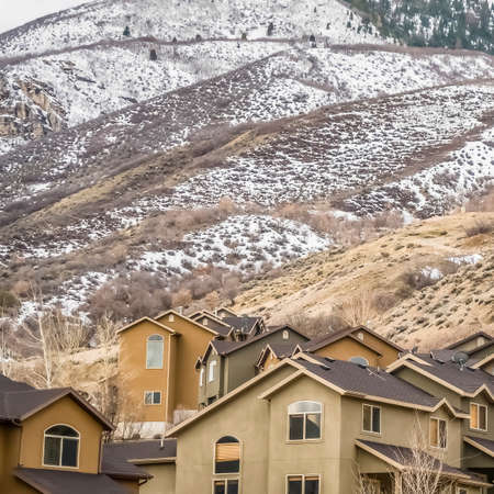 Square Exterior view of homes with snow topped mountain and cloudy sky background. The houses have pitched roofs, shiny windows, and brown walls.の写真素材