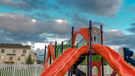 Panorama Playground with slides swings table and seat against a picket fence and houses. Over the landscape is a vast blue sky with puffy gray clouds.の写真素材