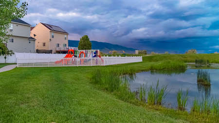 Panorama frame Bench and pond on a grassy park with playground and homes in the background. Towering...