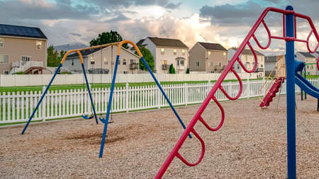 Panorama frame Climbing bars swings and slides on a neighborhood playground against cloudy sky. Vast grassy terrain, houses, and mountain can be seen in the background.の写真素材