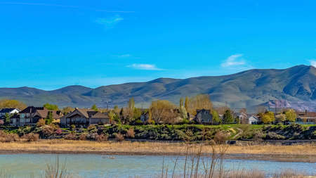 Panorama frame View of a lake with beautiful houses near the shore with brown grasses. A mountain...