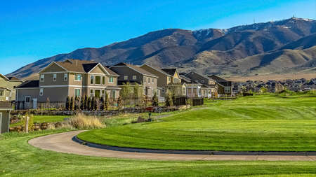 Panorama frame Narrow paved road curving through a grassy field in front of a residential area. A towering mountain under clear blue sky can be seen in the background.の写真素材