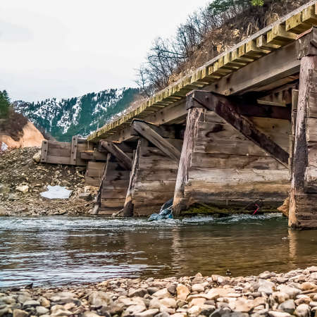 Square frame Rustic wooden bridge over a rocky stream with shiny water. In the background is a view...