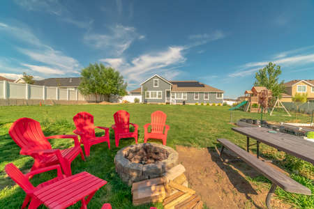 Red outdoor chairs and picnic table with bench around a circular stone fire pit. A playground can...