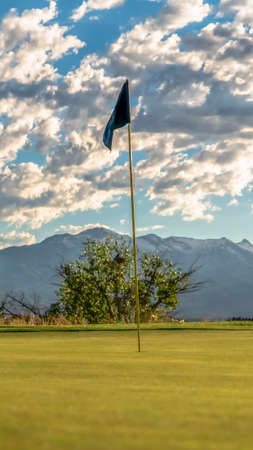 Vertical frame Focus on the flagstick of a golf course against trees mountain and cloudy sky. The yellow pole is inside a hole and has a black cloth at the tip.の写真素材