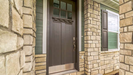 Panorama frame Home entrance with a glass paned brown wooden door and stone brick wall. The front window has a brown wooden shutter and white interior blinds.の写真素材