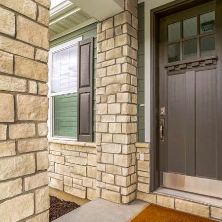 Square frame Home entrance with a glass paned brown wooden door and stone brick wall. The front window has a brown wooden shutter and white interior blinds.の写真素材