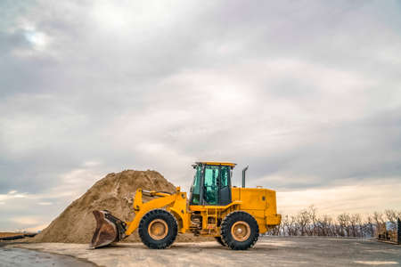Bulldozer in front of a mound of soil with leafless trees and cloudy sky background. The yellow...