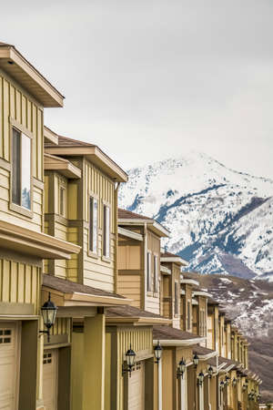 Row of townhouses in winter with snow covered mountain and cloudy sky background. The residences have white wall sidings, sliding windows, and garage doors with outdoor lamps.の写真素材