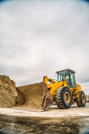 Front of a yellow bulldozer with mound of soil and cloudy sky in the background. The construction equipment has a dirty metal bucket and black rubber wheels.の写真素材