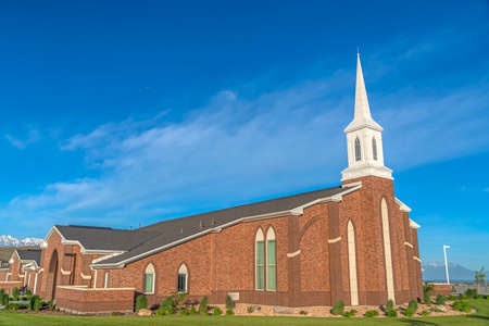 Exterior of a church with red brick wall arched windows and white steeple. Snow peaked mountain and blue sky can be seen in the background on this sunny day.の写真素材