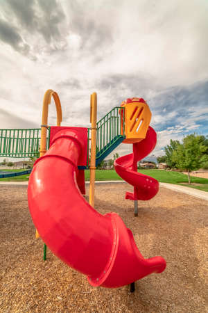 Red closed tube slide and spiral slide at a playground against cloudy blue sky. Vivid green grasses...